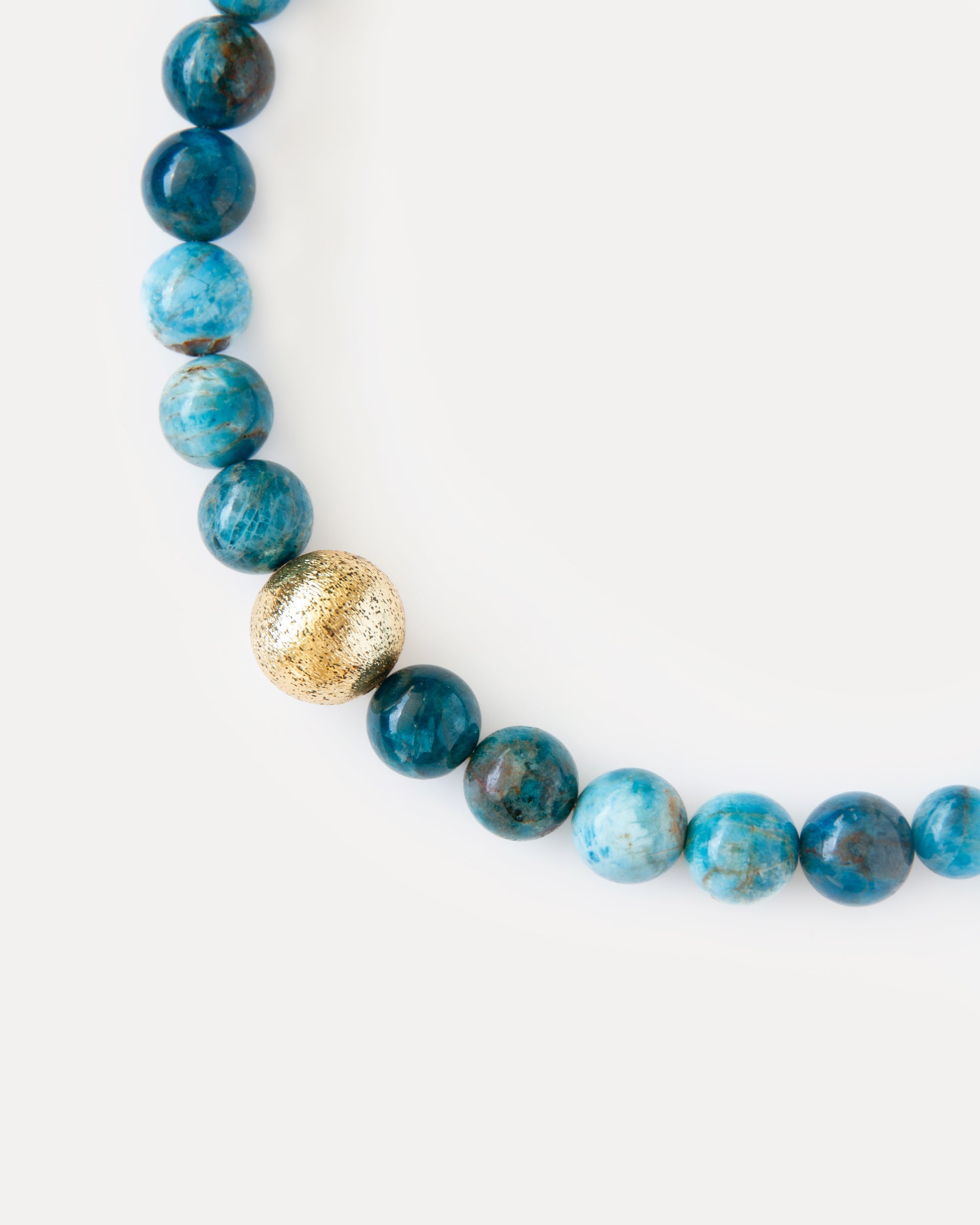 Ocean View Necklace 
An exquisite close-up focusing on the textured 18K gold-plated brass bead beautifully nestled among the smooth, deep blue apatite stones. This detailed shot emphasizes the contemporary design and the vibrant, rich hues of the natural apatite. It highlights the intriguing play of light on the golden texture and the profound depth of color in the natural stone, creating a refined and fashionable accent for any personal style.