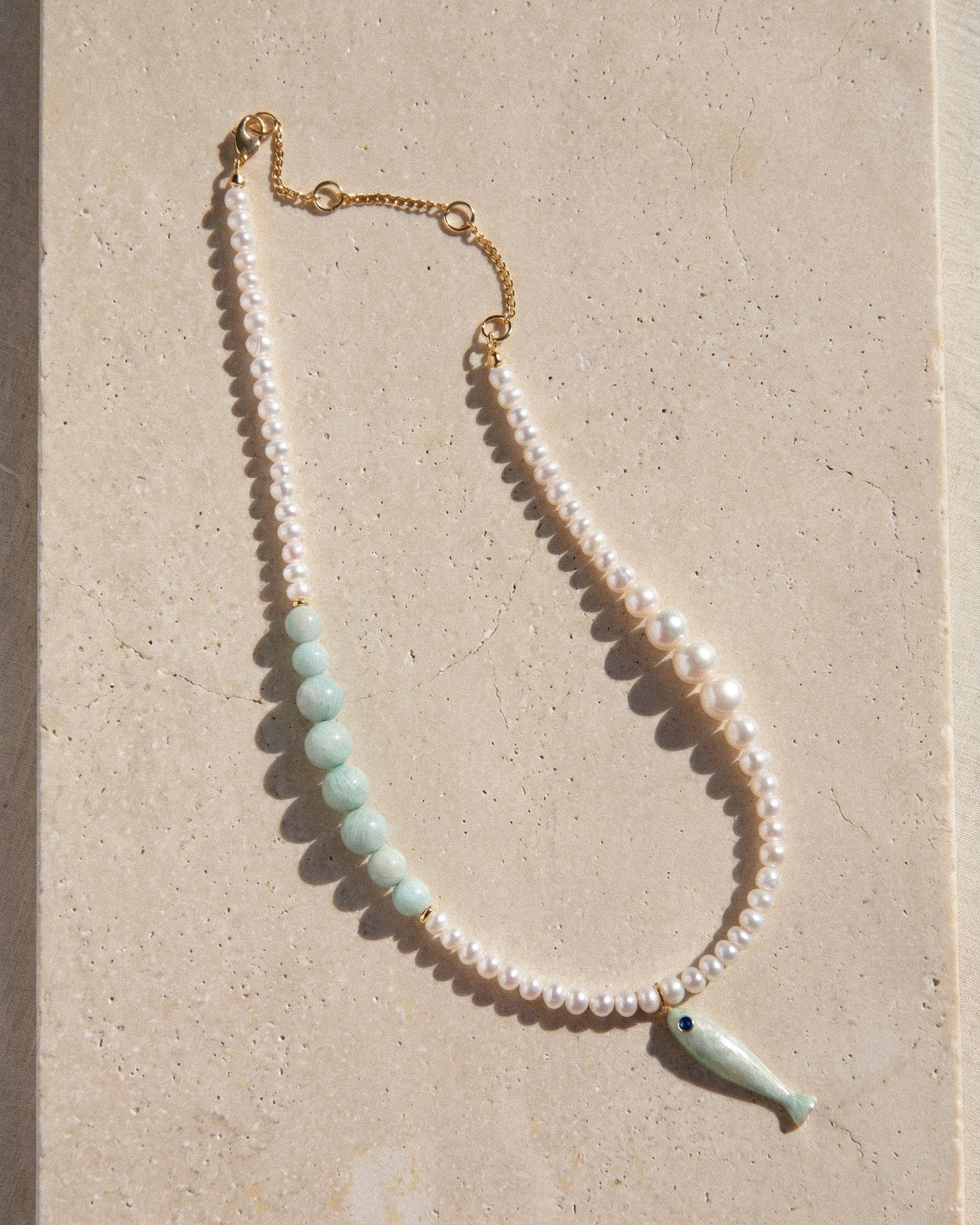 freshwater pearl necklace with pale amazonite beads and ceramic fish pendant styled on warm stone surface