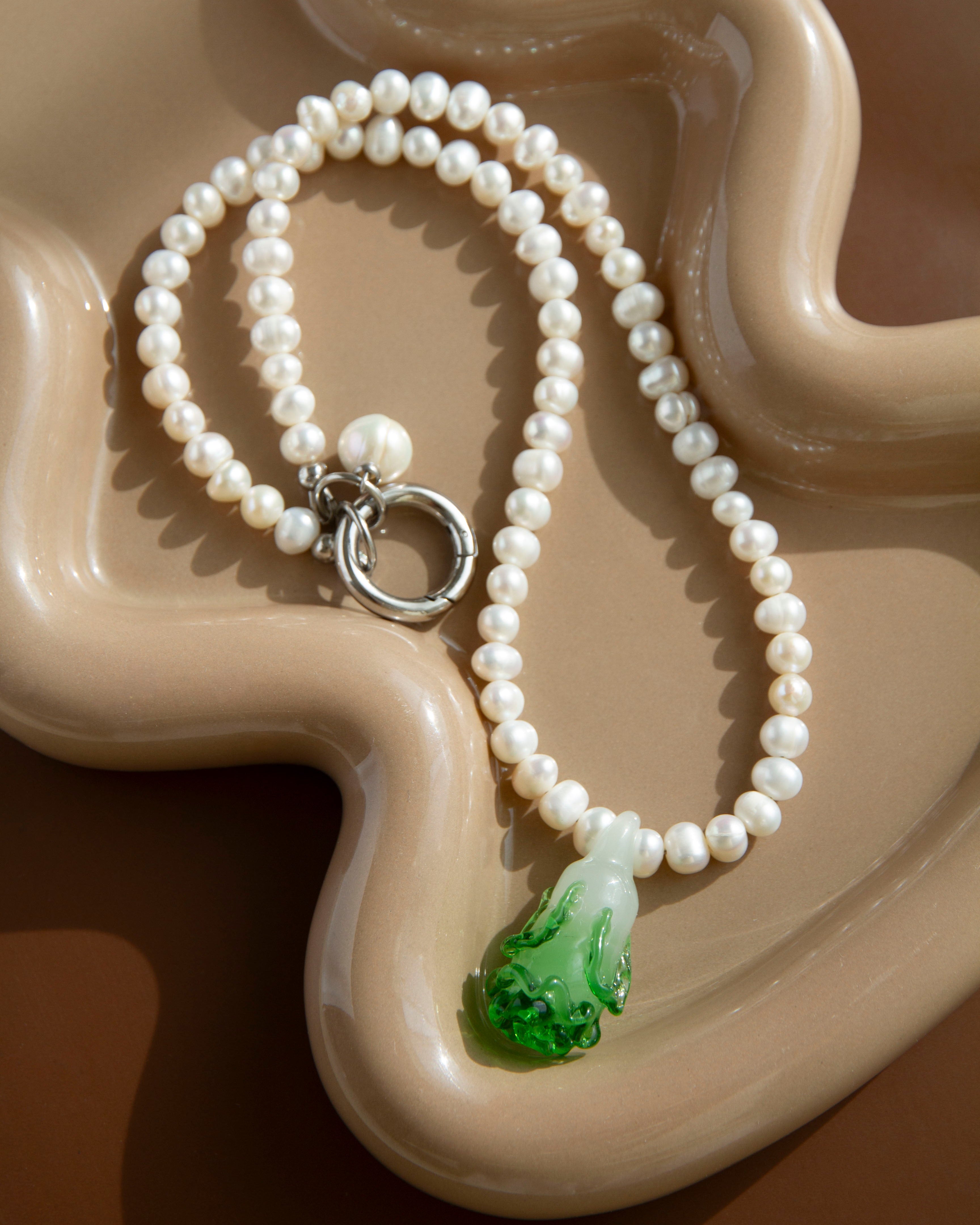 Cabbage Pearl Necklace on a beige ceramic dish – strand of freshwater pearls with green lampwork bok choy pendant and steel clasp.