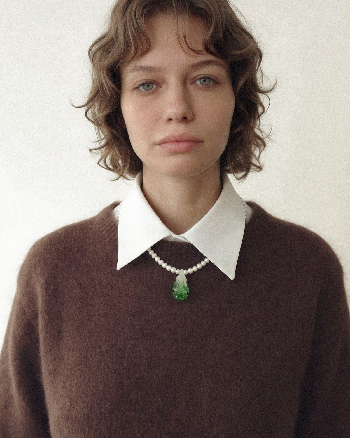 Model wearing a pearl necklace with a green glass cabbage pendant below a white shirt collar, over a brown sweater.