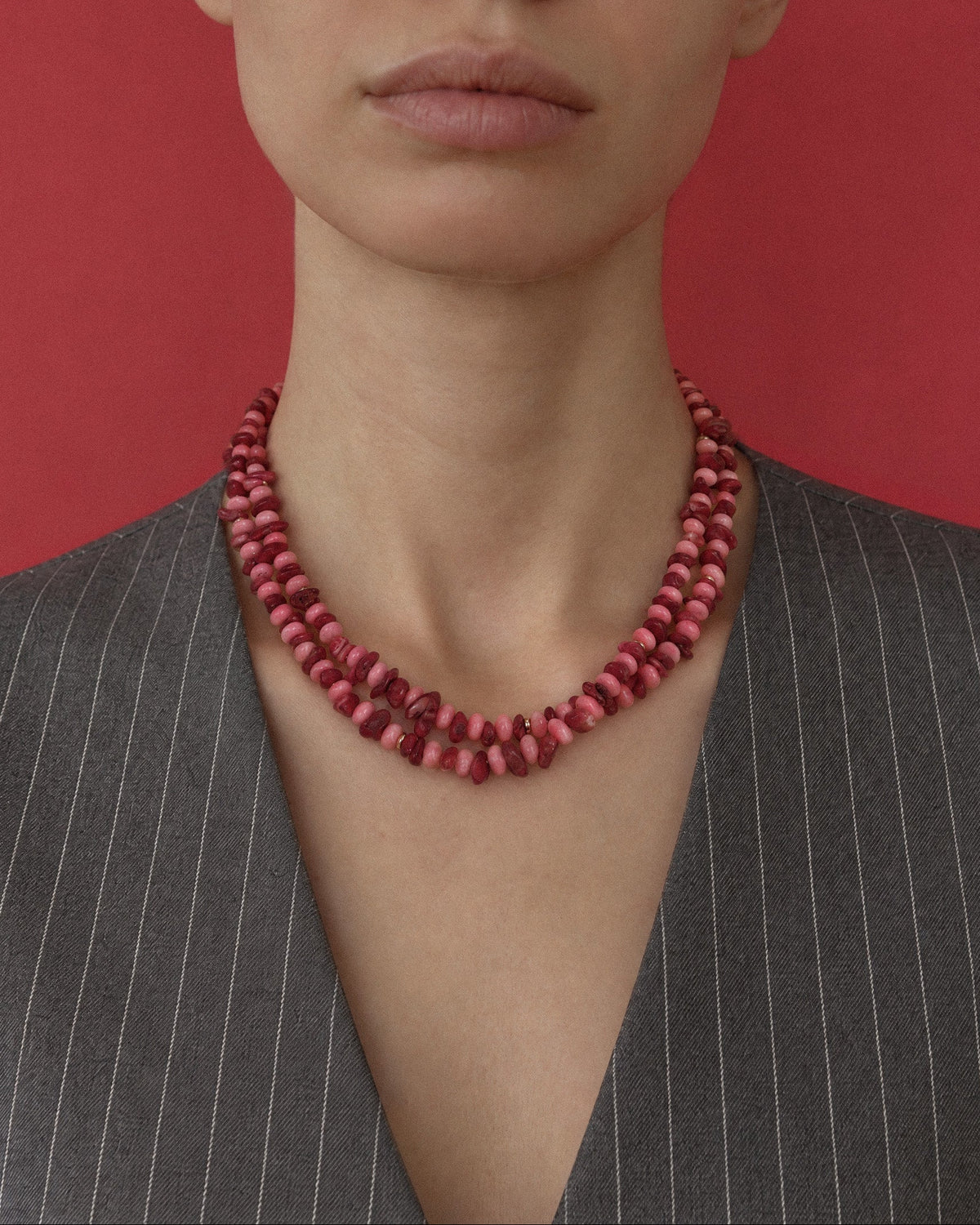 A close-up of a model wearing a chunky, multi-strand pink and red coral beaded necklace, paired with a gray pinstripe blazer against a solid red background.