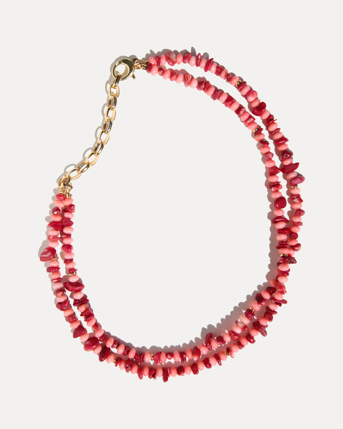A double-strand necklace made of small, irregular pink and red coral beads with a gold-tone chain extender and lobster clasp, displayed on a light gray background.