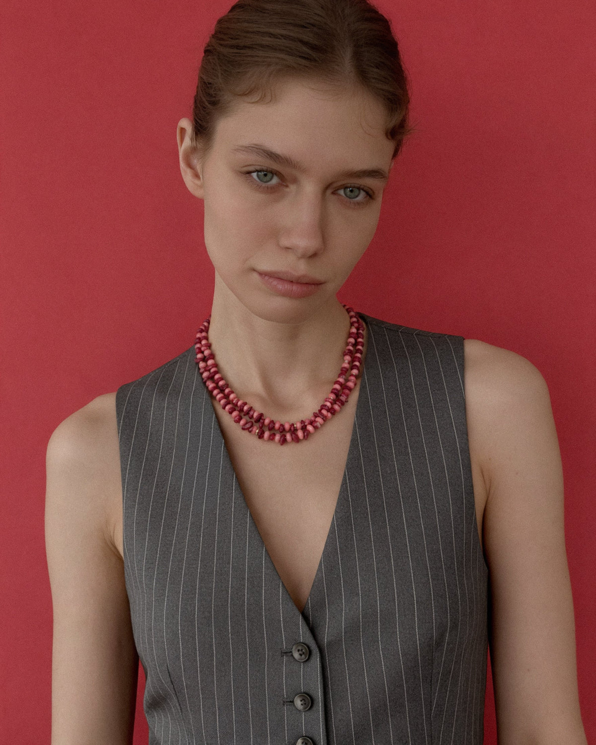 A model with her hair pulled back wears a gray pinstripe vest and a chunky pink and red coral necklace with gold details set against a solid red background.