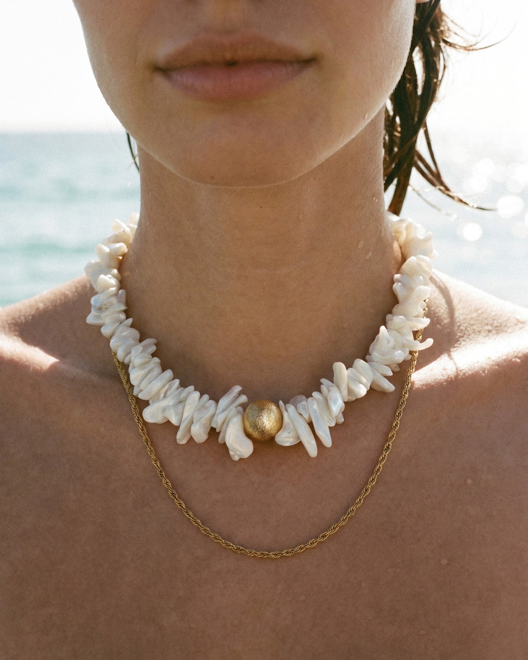 statement shell choker necklace with white shell beads and layered gold chain, close-up on model by the sea