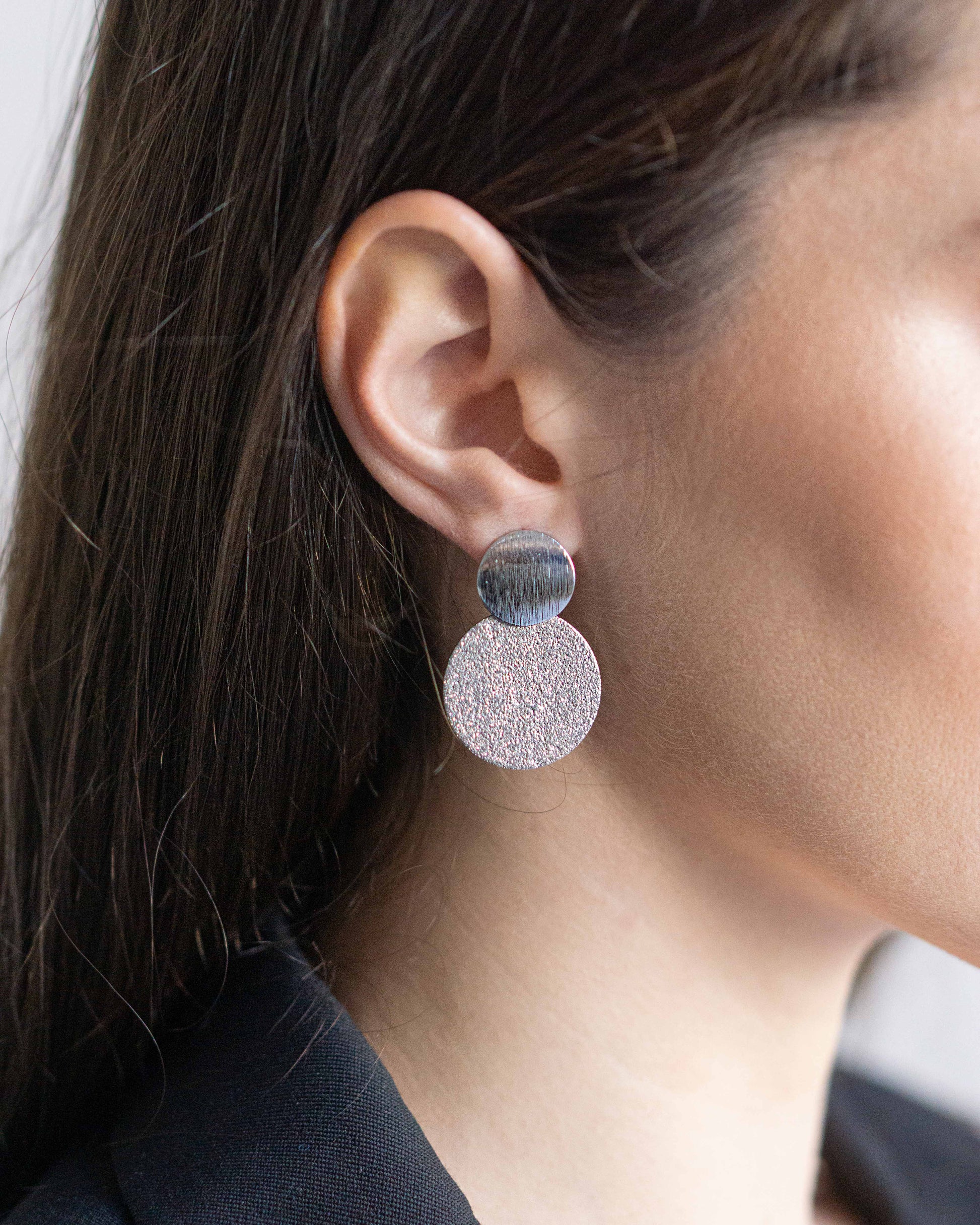 Close-up of silver double-disc statement earrings with soft matte and textured finish, adding a sculptural, modern accent to minimalist everyday looks.