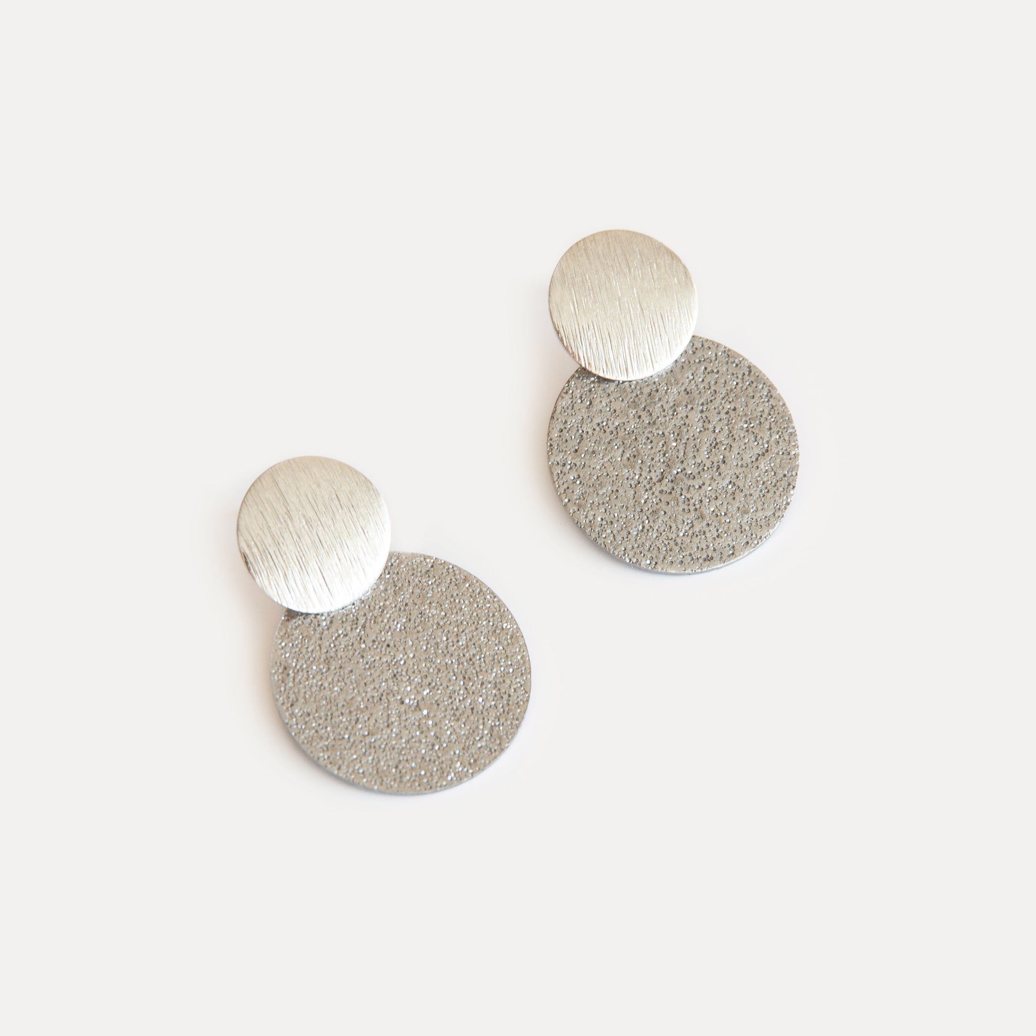 Minimalist silver statement earrings with two textured discs in contrasting finishes, crafted from stainless steel for a modern, sculptural look.