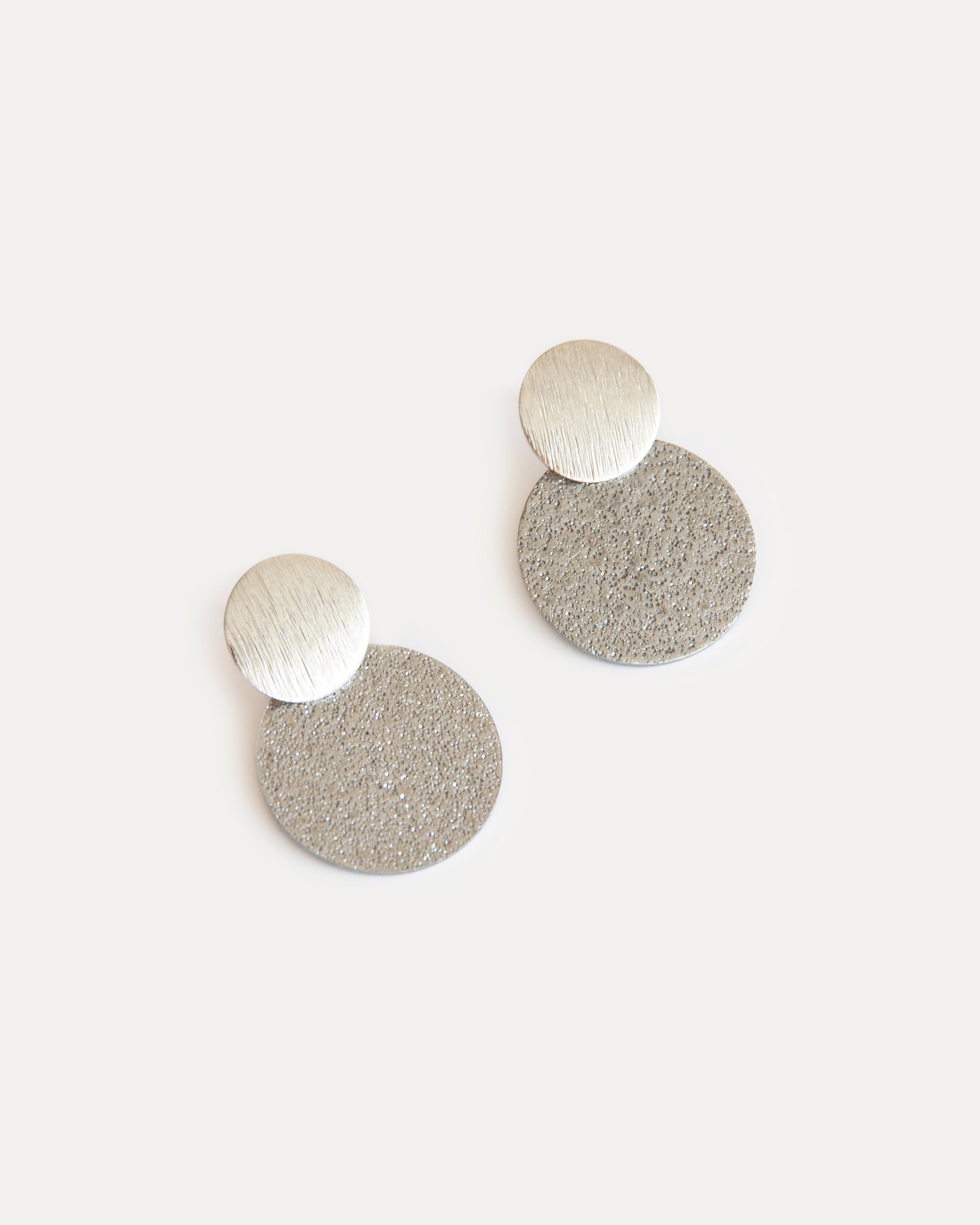 Minimalist silver statement earrings with two textured discs in contrasting finishes, crafted from stainless steel for a modern, sculptural look.