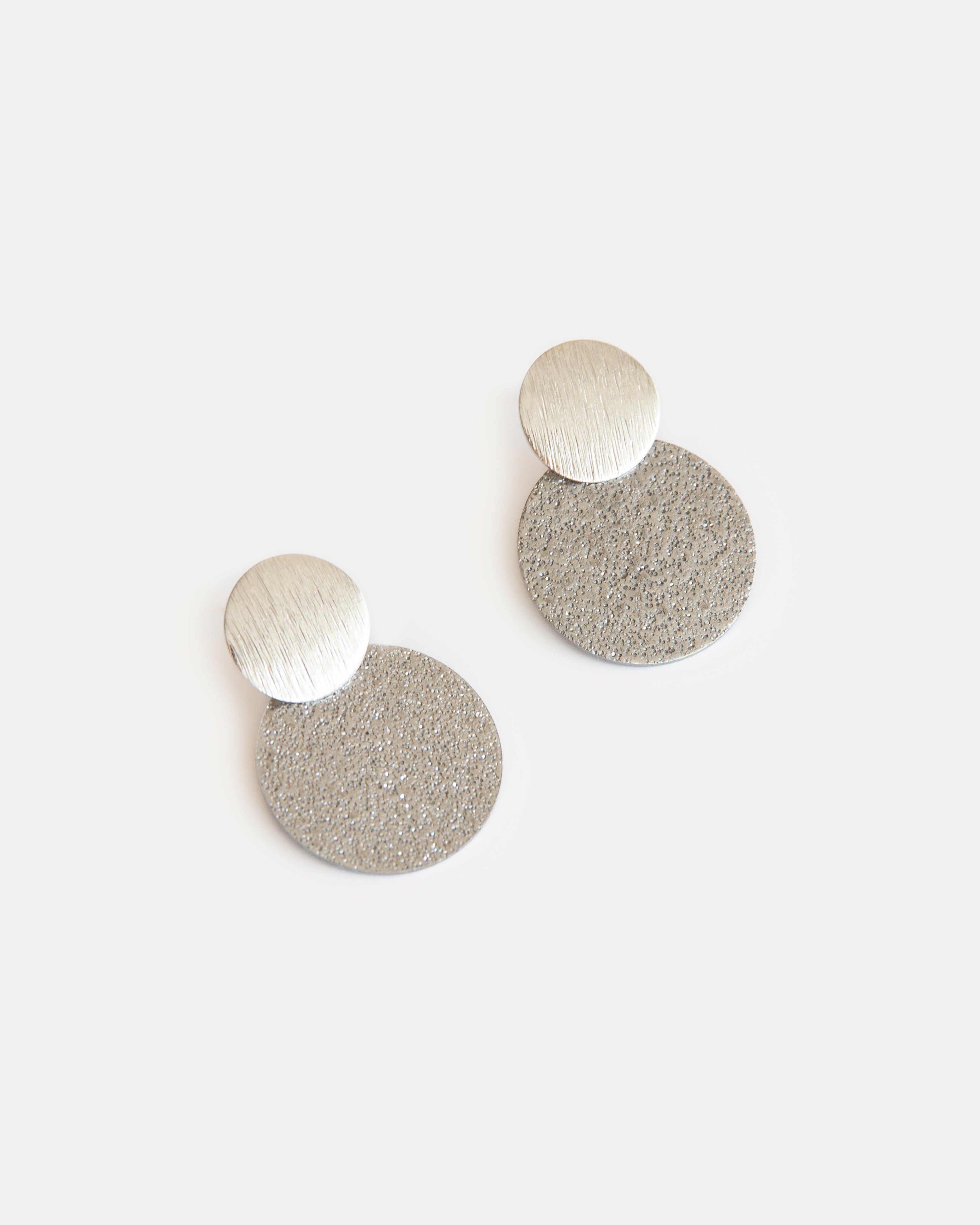 Minimalist silver statement earrings with two textured discs in contrasting finishes, crafted from stainless steel for a modern, sculptural look.