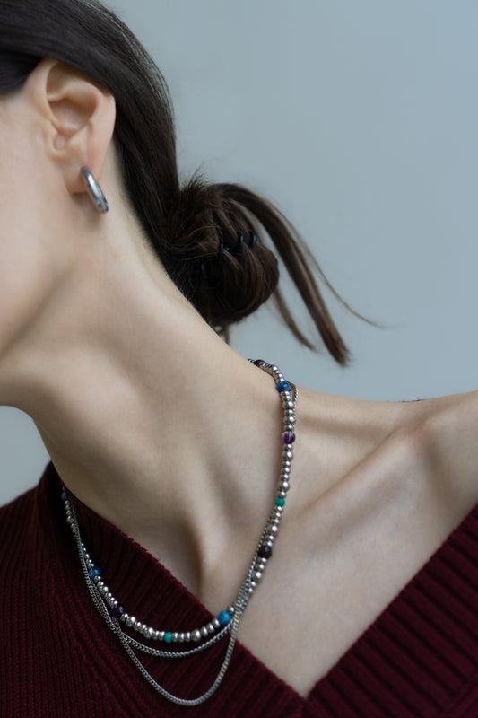 Close-up of a model's ear, neck, and shoulder, showcasing the elegant layering of the GEMS necklace with its vibrant gemstone beads (amethyst, kyanite, garnet) and polished steel chains. A minimalist silver hoop earring completes the look. This image highlights the versatility and contemporary style of Jewell Reef's handmade jewelry, perfect for enhancing a sophisticated profile and adding a touch of color to any outfit