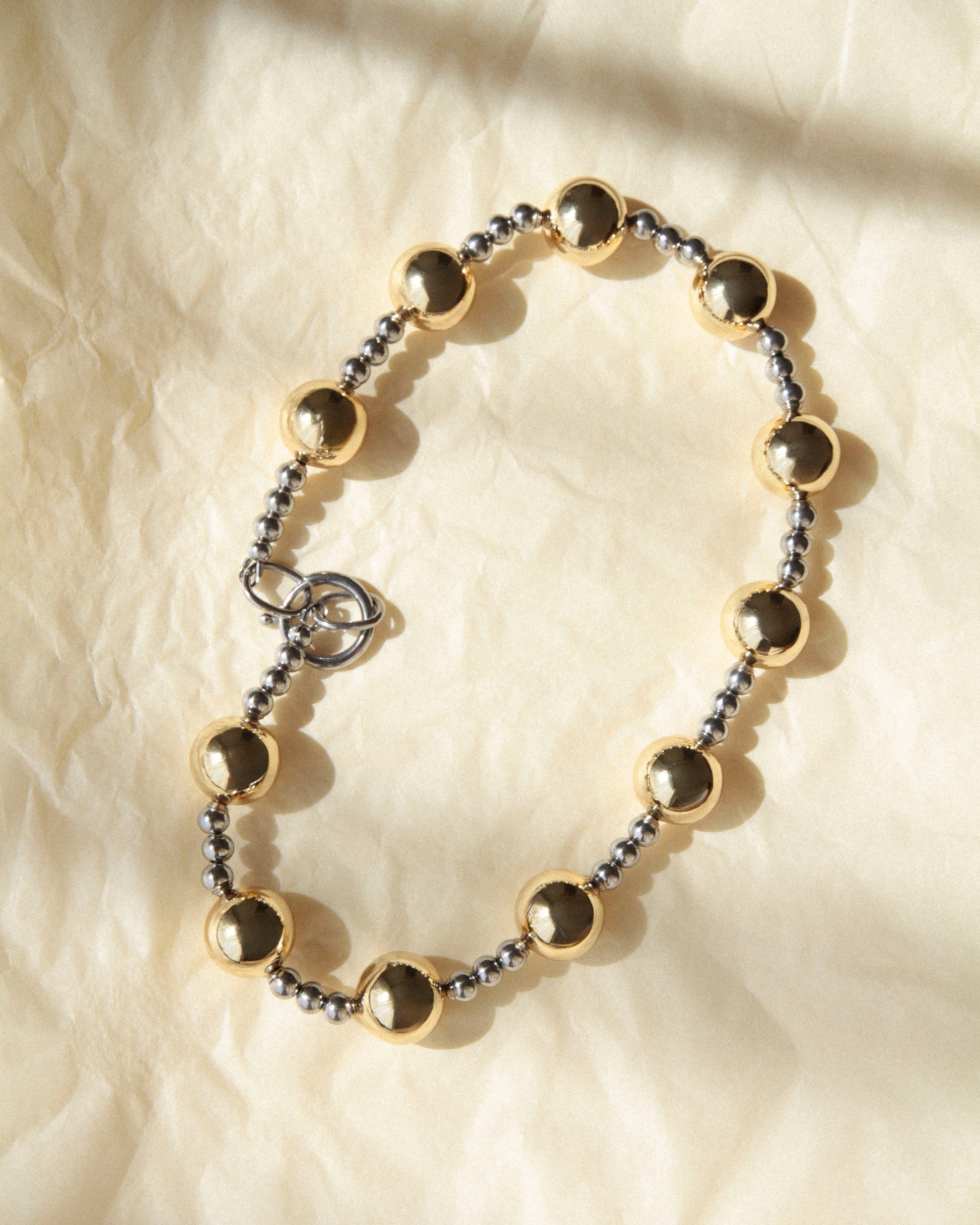 mixed metal statement necklace with gold and silver beads on soft fabric, natural light creating gentle shadows and subtle reflections