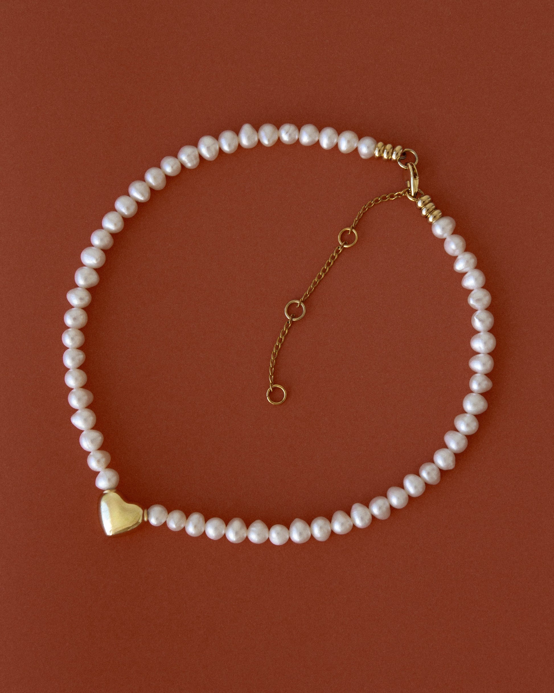 Heart of Gold pearl necklace with 18K gold-plated heart pendant on a warm terracotta background — elegant, timeless jewelry with soft contrast and glow.