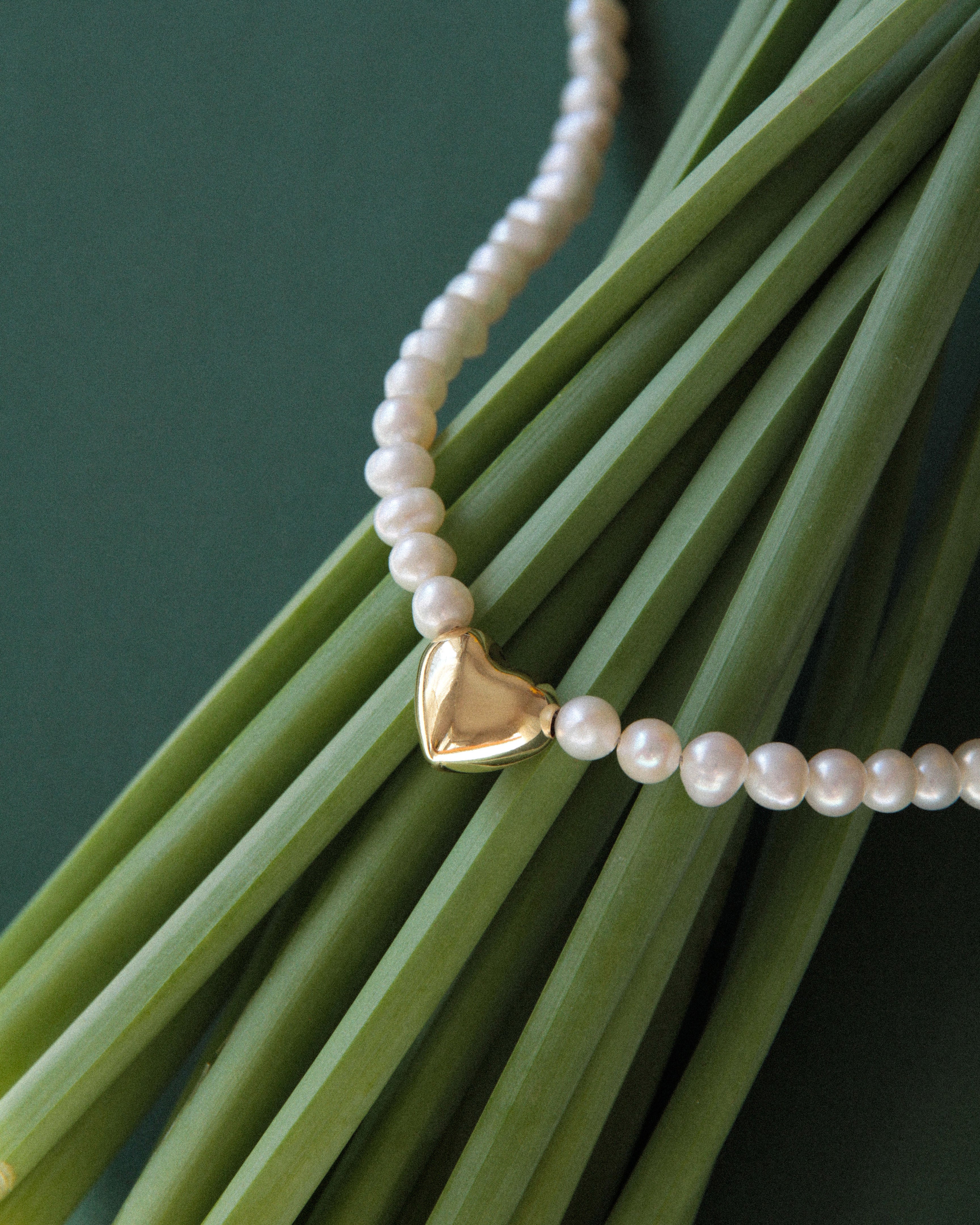 Freshwater pearl choker necklace detail with gold heart charm, close up of pearl texture and polished pendant on green stems