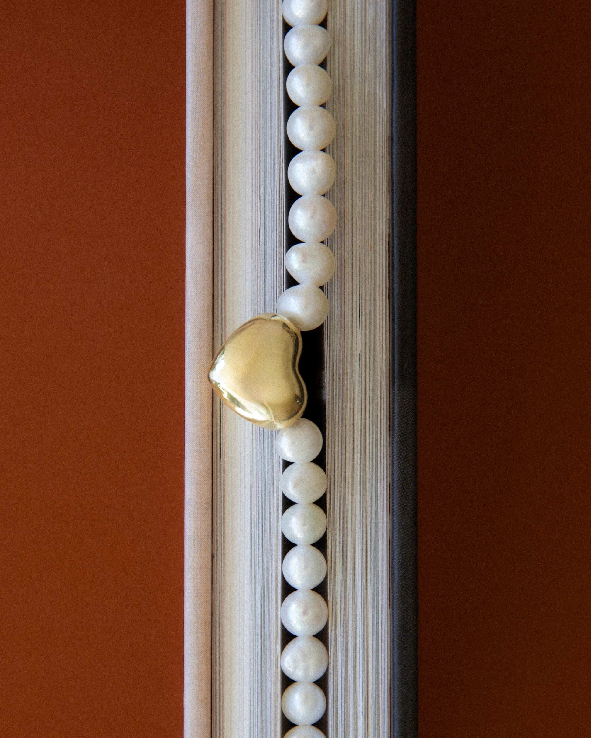Freshwater pearl necklace with gold heart charm styled between book pages, editorial jewelry composition with warm tones and soft texture