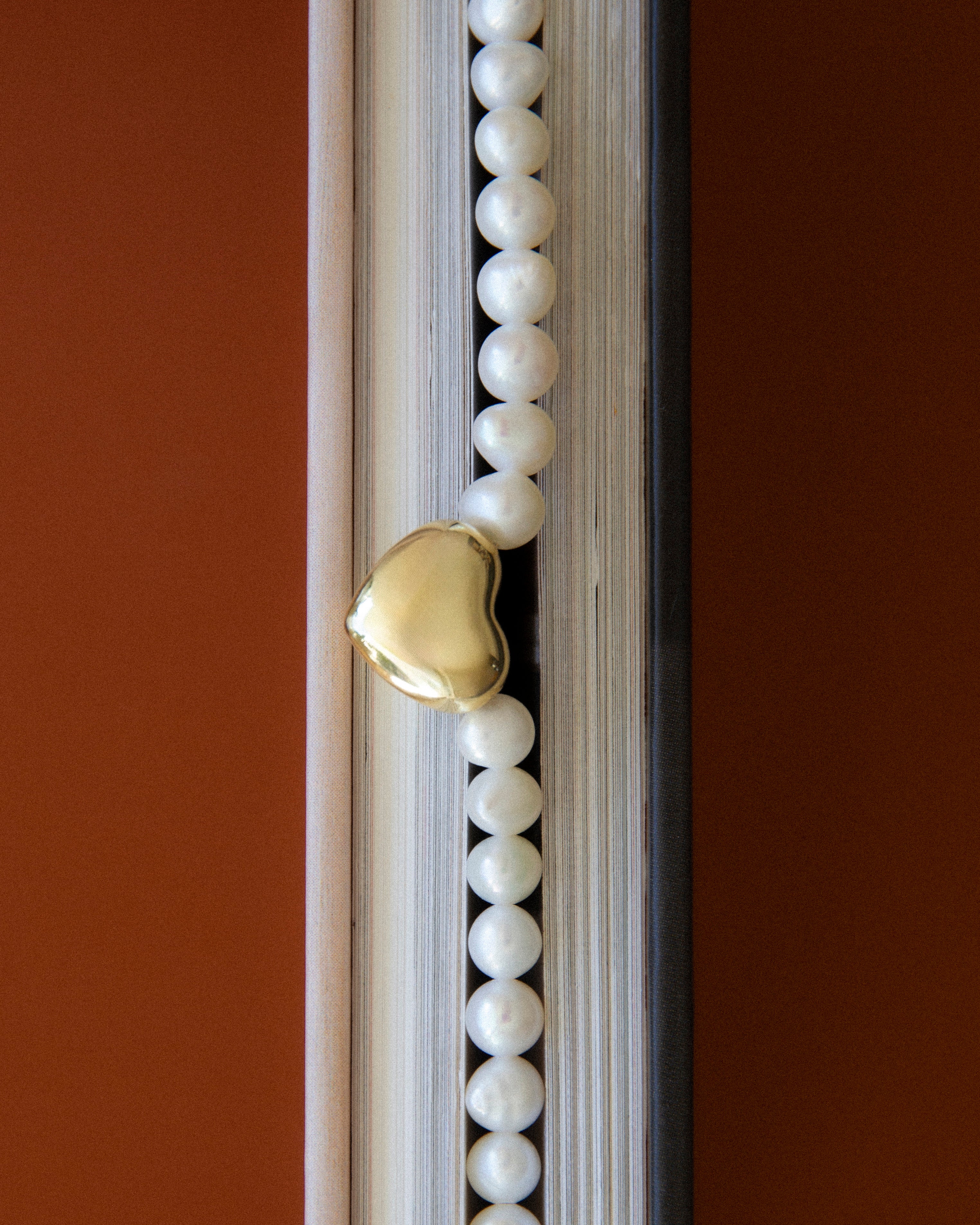 Freshwater pearl necklace with gold heart charm styled between book pages, editorial jewelry composition with warm tones and soft texture