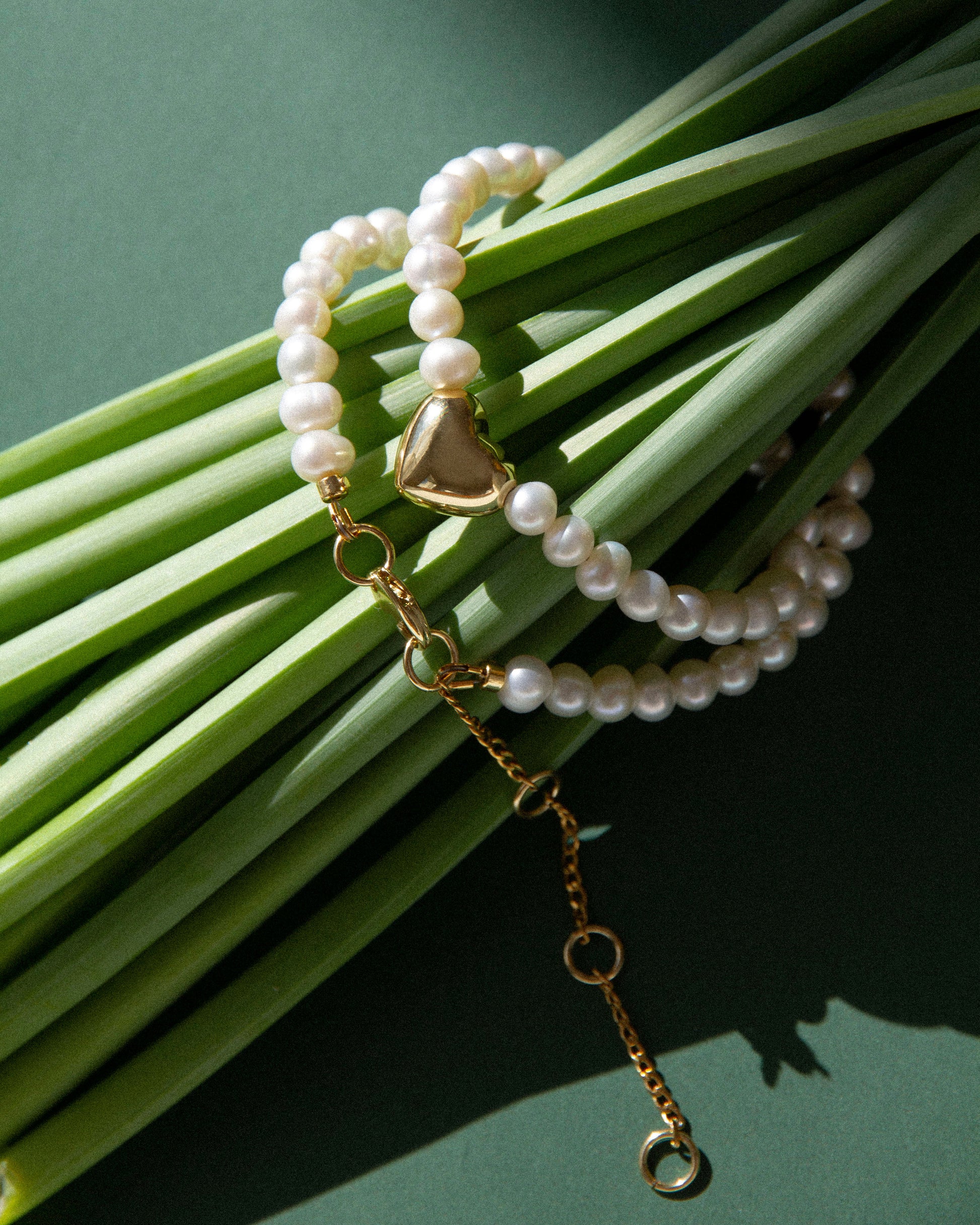 Freshwater pearl necklace with gold heart charm styled on green leaves, natural light jewelry styling with organic textures and soft contrast