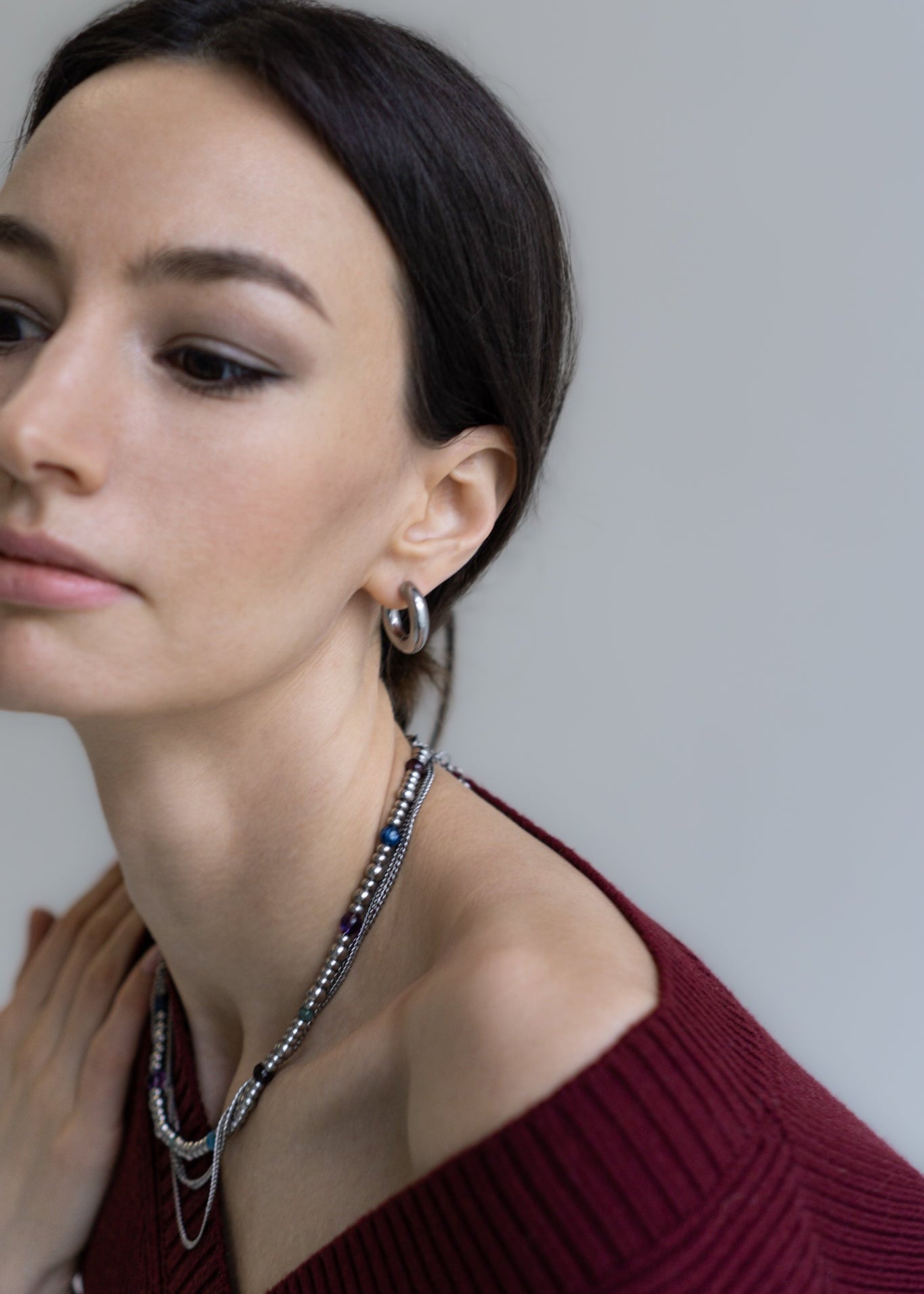 Close-up of a model showcasing the GEMS necklace, highlighting its vibrant gemstone beads (garnet, amethyst, kyanite, amazonite, apatite) and layered steel chains against a dark red top. This image emphasizes the necklace's stylish impact on a woman's décolletage, paired with minimalist silver hoop earrings. Perfect for capturing the modern elegance and versatile appeal of Jewell Reef's unique jewelry, ideal for elevating everyday looks and special occasions.