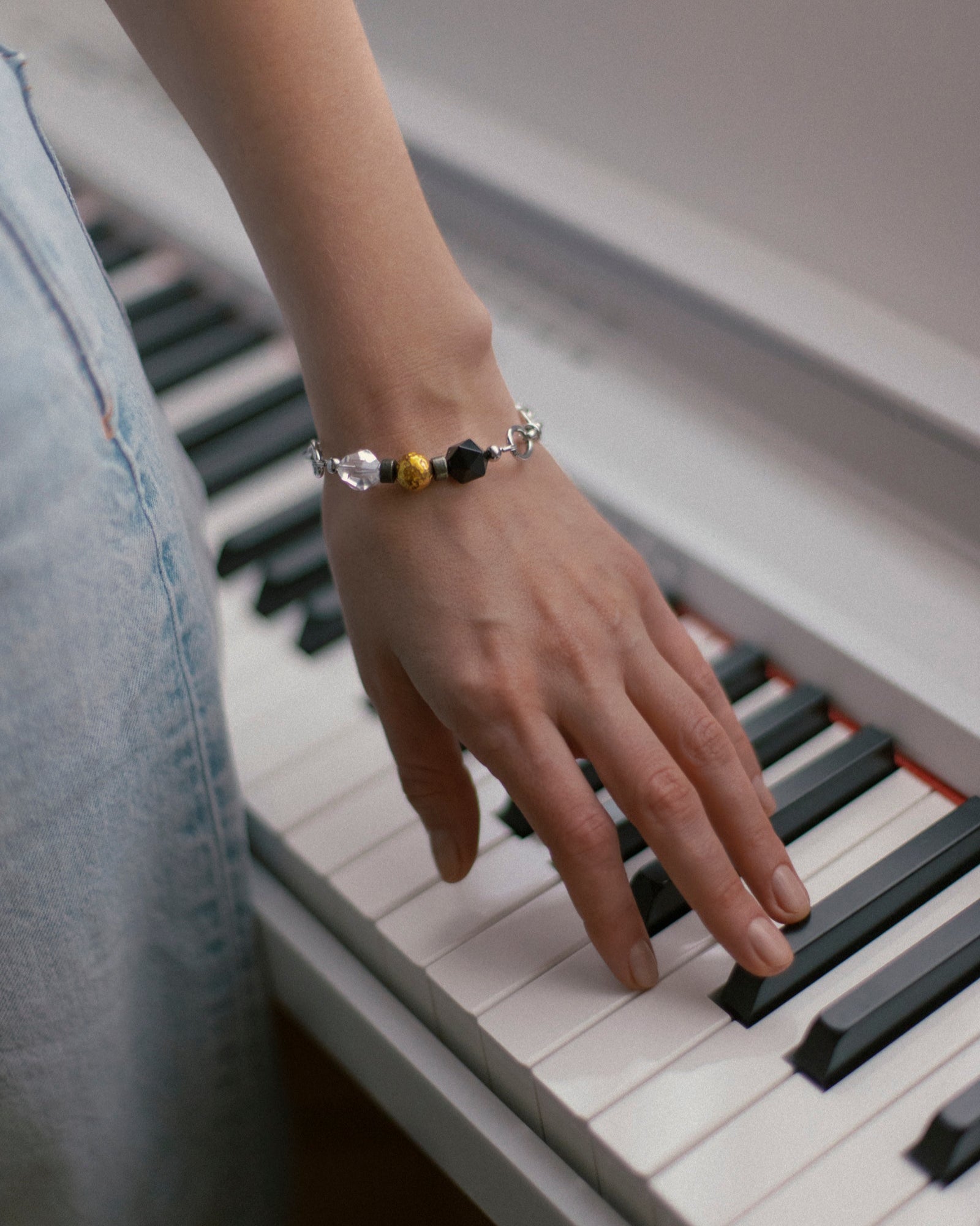 Woman's hand resting gracefully on piano keys, highlighting the Jazz Bracelet on her wrist. The bracelet's design, featuring clear quartz, black agate, and 18K gold-plated brass beads on a stainless steel chain, beautifully complements the musical theme. This image showcases Jewell Reef's unique, minimalist jewelry, perfect for daily wear, expressing rhythm and modern style with intention.
