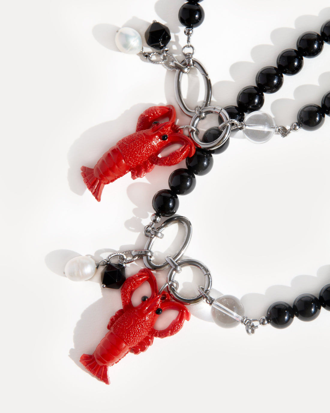 Lobster Noir Necklace by Jewell Reef — bold statement jewelry handcrafted with polished black agate beads, clear quartz, and stainless steel. The photo shows two versions of the necklace with detachable pendants: a vivid red lobster charm and a duo of faceted black agate with a freshwater pearl. Playful ocean-inspired necklace with versatile styling.