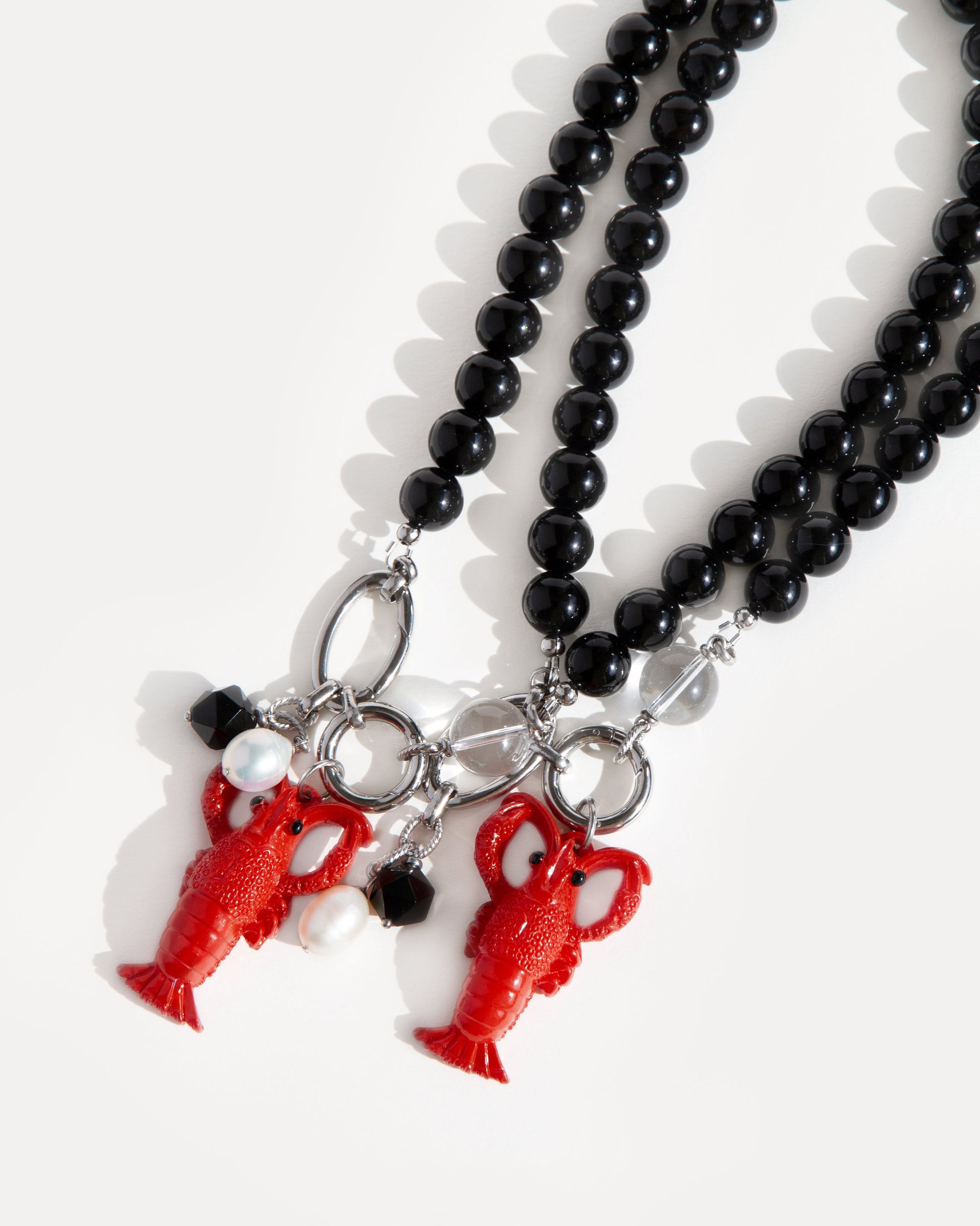 Two Jewell Reef Lobster Noir Necklaces displayed on white background — handcrafted statement jewelry made with polished black agate beads, stainless steel, and detachable pendants. Each necklace features a bold red lobster charm, faceted black agate, clear quartz, and a freshwater pearl. Playful ocean-inspired necklaces that combine fun and elegance, perfect for layering or wearing as a striking single statement piece.