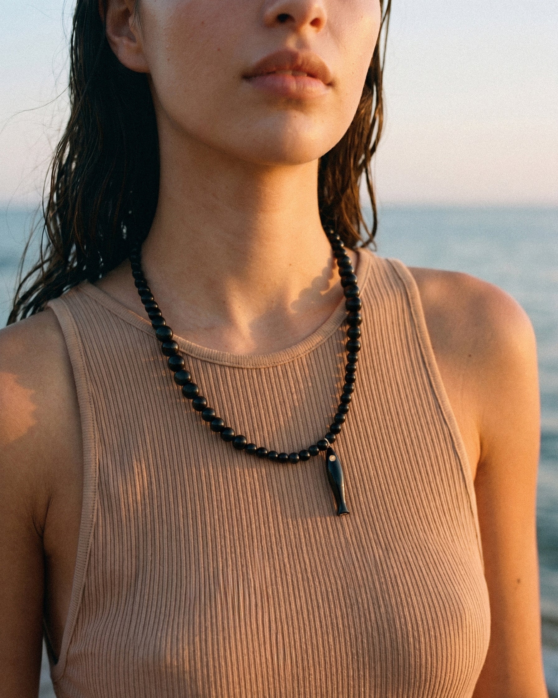 Close-up of model wearing black agate necklace with ceramic fish pendant and gold details by the sea at sunset