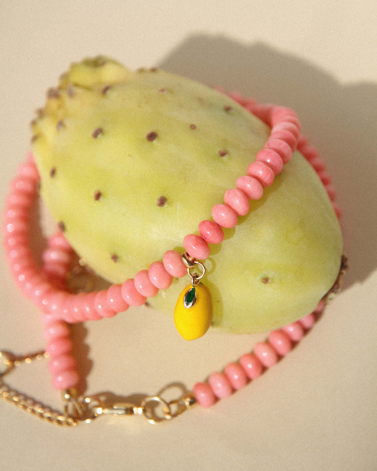Pink coral necklace with an enamel lemon charm styled over a yellow cactus fruit in a sunlit still life.