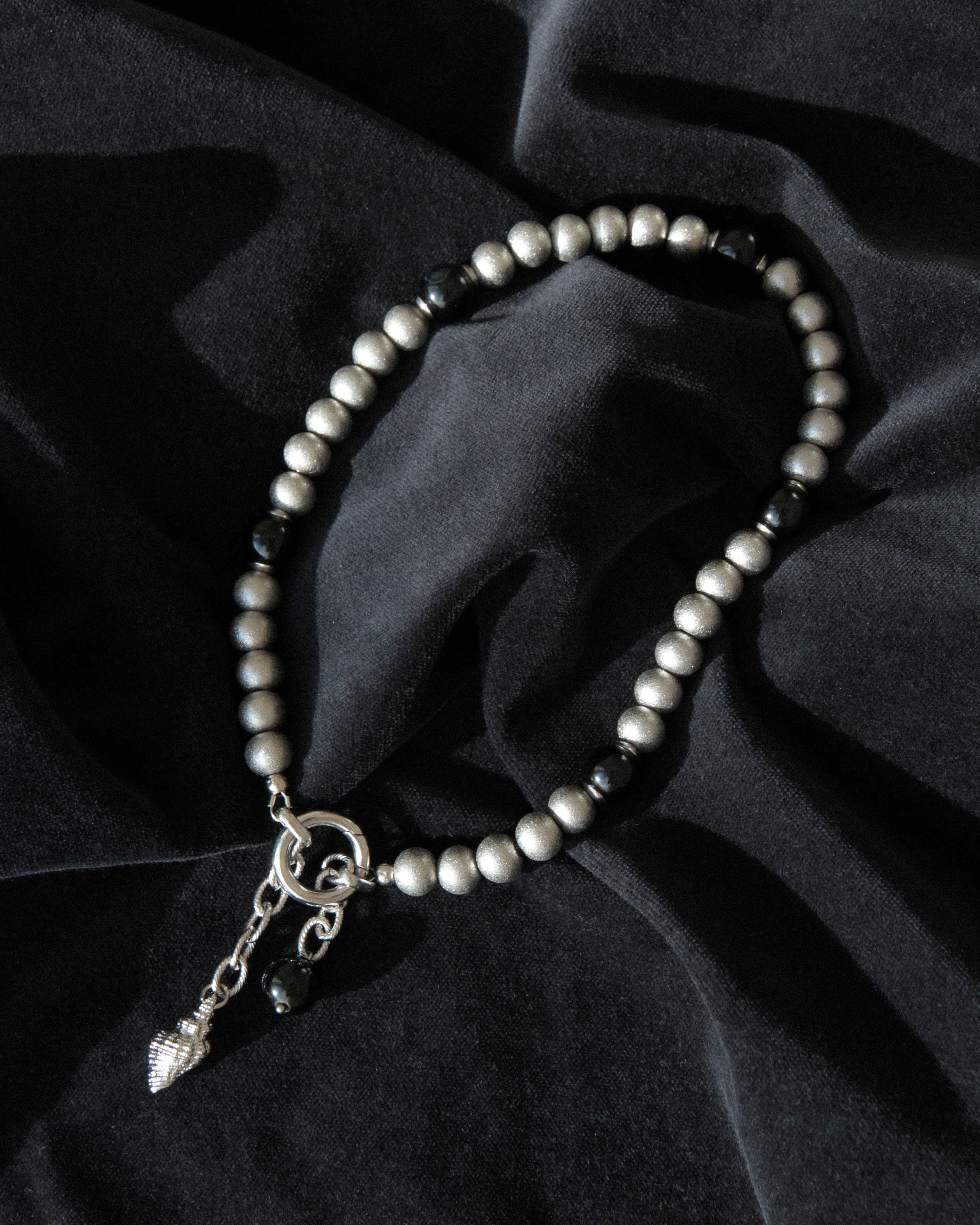 Black agate and stainless steel beaded choker necklace with front ring clasp and shell drop pendant arranged on dark velvet fabric