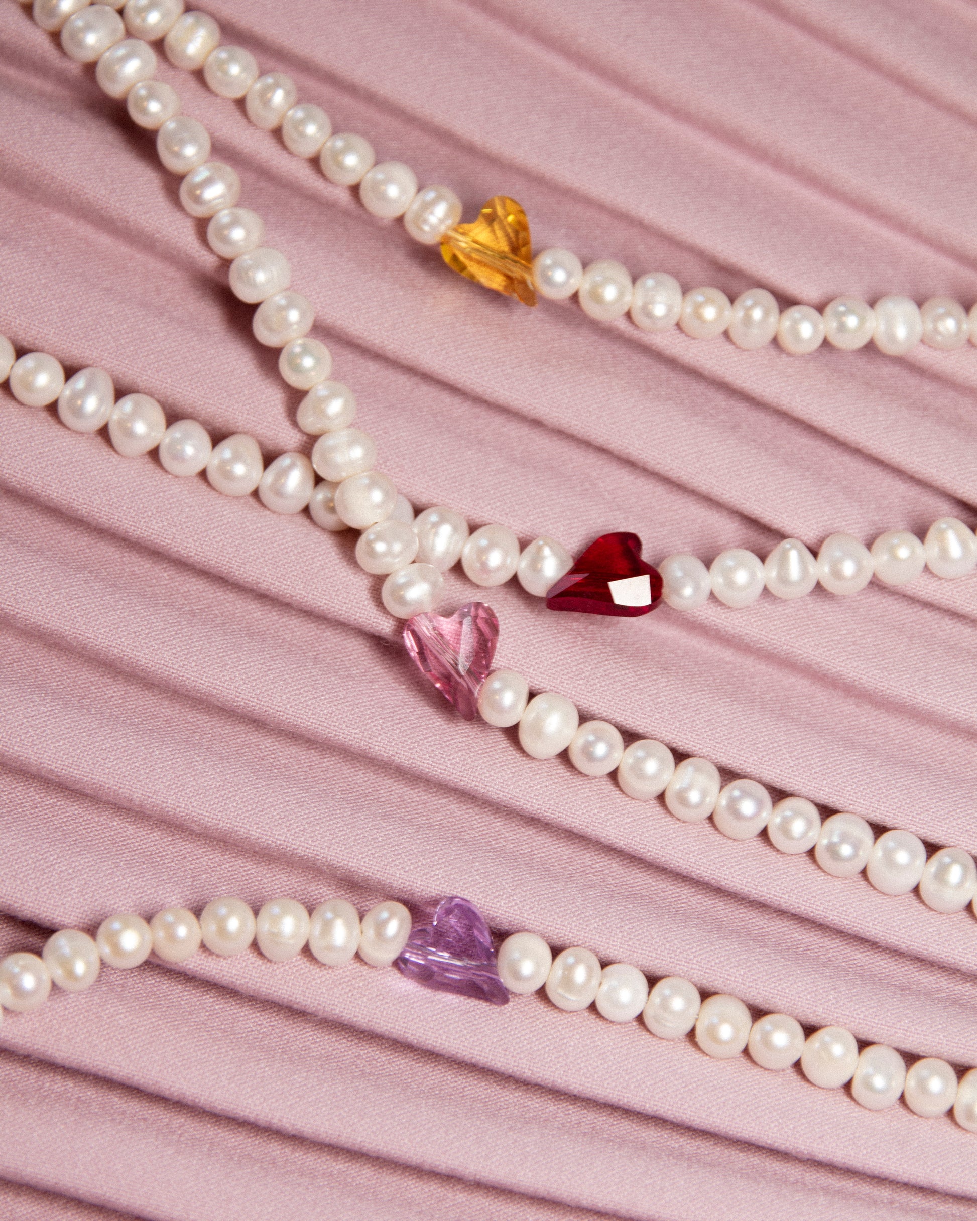 Flatlay of Sweetheart pearl choker necklace with crystal hearts in all colors — yellow, pink, red, and purple — styled on soft fabric background.