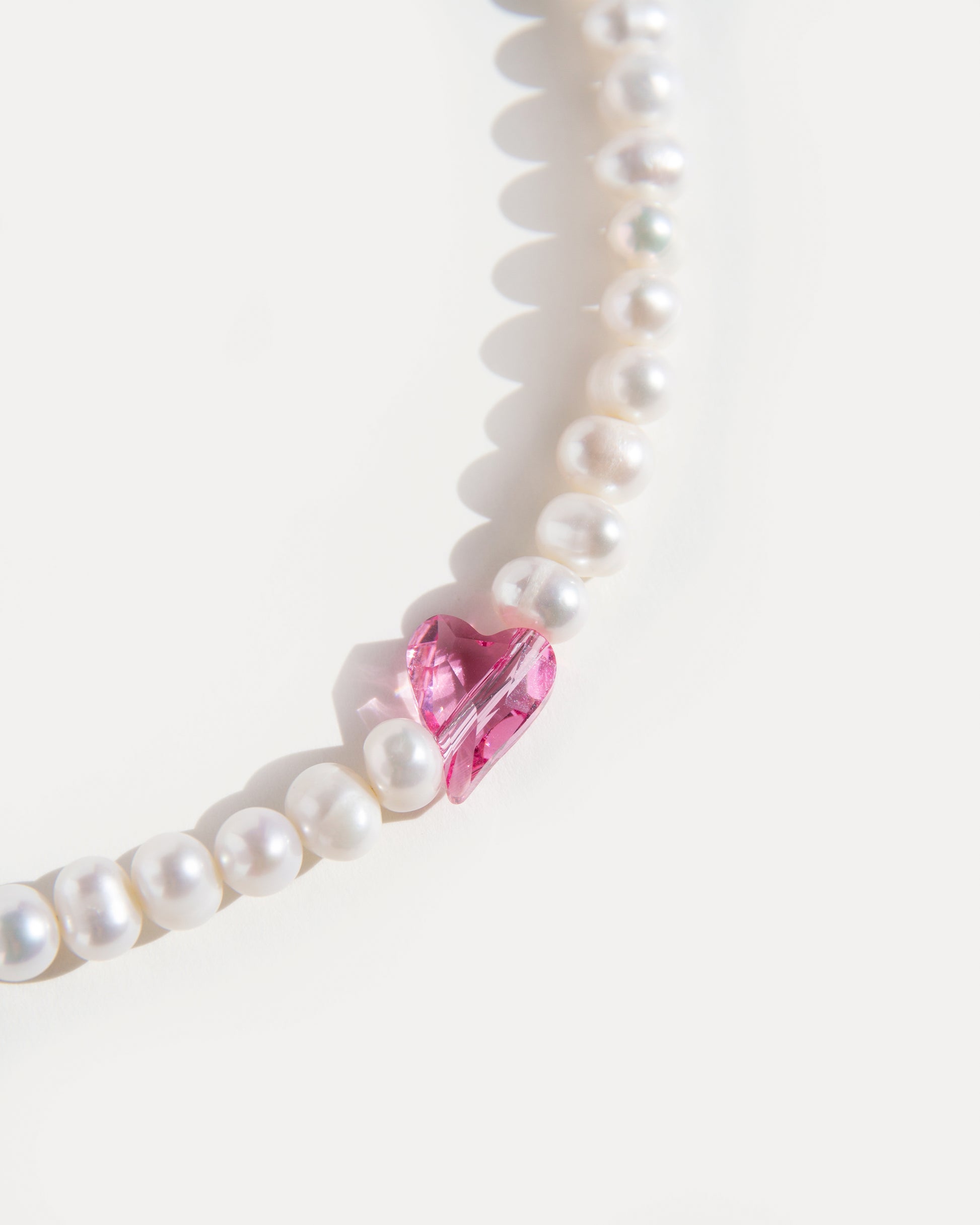 Sweetheart pearl choker necklace close-up showing natural pearls with a pink crystal heart accent, feminine and playful jewelry detail.